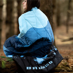 Person sitting on a log wearing a blue and black jacket with 'BRONCO' branding in a forest setting.